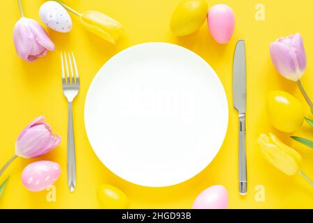 Easter table setting. White plate, decorative easter eggs, tulips and silver cutlery on yellow background Stock Photo