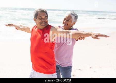 Happy biracial senior woman helping man practice yoga with arms outstretched at sunny beach Stock Photo