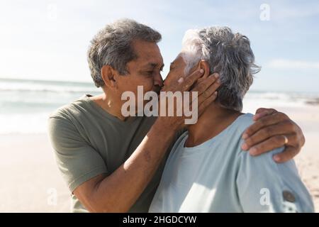 Loving biracial senior man kissing woman on lips at beach during sunny day Stock Photo