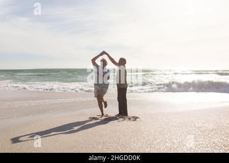 Full length of happy retired senior multiracial couple dancing on shore at beach during sunny day Stock Photo