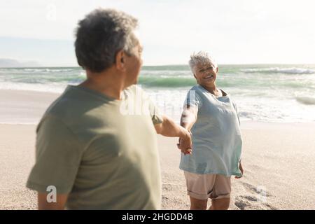 Happy biracial senior woman holding hand of man while looking at him on sunny beach Stock Photo