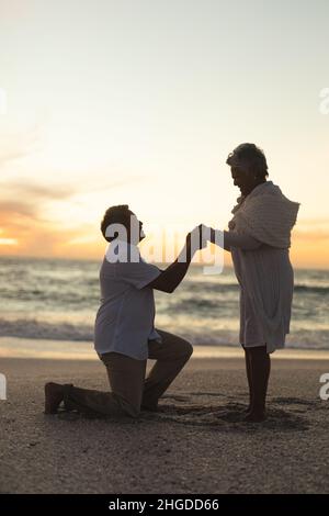 Side view of senior biracial man holding hands with woman while kneeling and proposing at beach Stock Photo