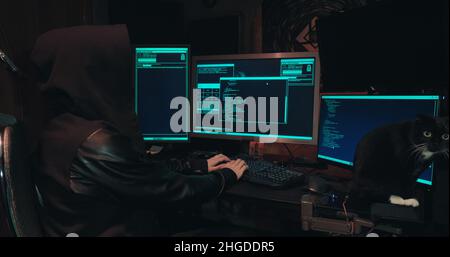 Hacker in a hood sits in front of computer screens and hacks databases Stock Photo