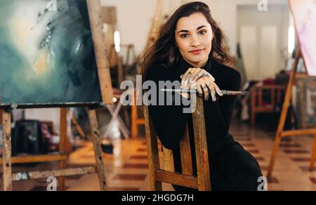 Young female painter looking at the camera confidently while sitting on a chair in her workshop. Self-employed female artist holding a paintbrush with Stock Photo