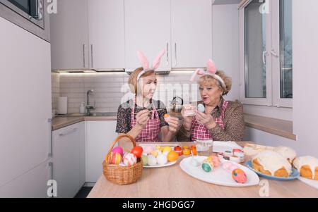 two happy elderly women with rabbit ears on their heads are painting Easter eggs at home in the kitchen. Stock Photo
