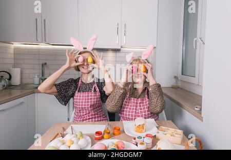two happy elderly women with rabbit ears on their heads are holding Easter eggs near their eyes and laughing merrily. Stock Photo