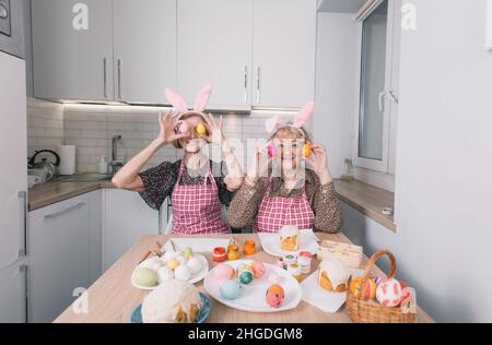 two happy elderly women with rabbit ears on their heads are holding Easter eggs near their eyes and laughing merrily. Stock Photo