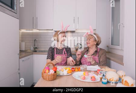 two happy elderly women with rabbit ears on their heads are painting Easter eggs at home in the kitchen. Stock Photo