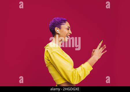 Wireless video calling. Happy young woman smiling cheerfully while holding a smartphone and wearing wireless earphones. Woman with purple hair taking Stock Photo
