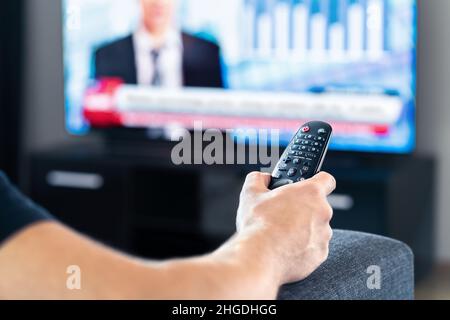 Man watching tv news program and sitting on couch home. Television remote control in hand. Politics, business or finance network channel. Stock Photo