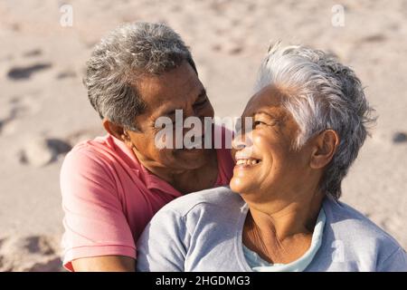 Loving retired multiracial senior couple looking at each other while sitting on sunny beach Stock Photo