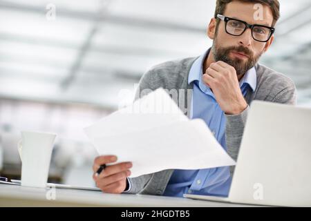 You'll need these documents. A young businessman going over paperowrk while working on his laptop in the office. Stock Photo