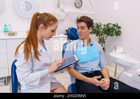 Redhead woman in medical uniform browsing tablet while sitting near male patient in modern dentist office Stock Photo