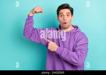 Photo of strong impressed young guy dressed violet sweatshirt pointing finger arms muscles big eyes isolated turquoise color background Stock Photo