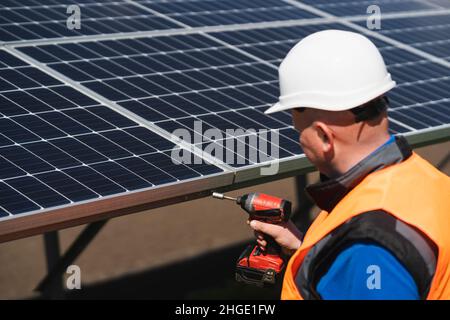Male electrical engineer in a hard hat with electric wrench maintains solar panels at power plant Stock Photo
