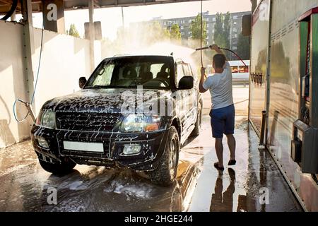 A man washes his car during the day, washes of the foam. A black SUV car at a self-service car wash. Washing the car with your own hands. Stock Photo
