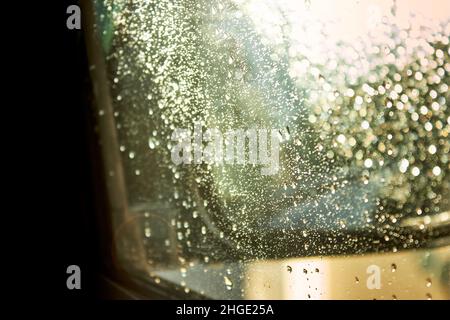 Water drops on the car window. The sunset sunlight shines on the glass. Sun glare. Abstract drip background in green-yellow color. Stock Photo