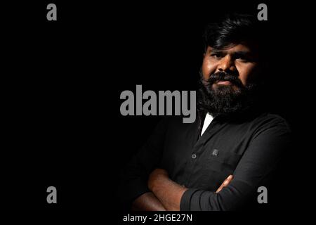 Young indain beardman with crossed arms standing against dark background - Conncept of self confident, positive emotion and happiness Stock Photo