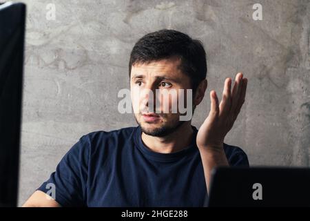 Handsome concentrated serious bearded man with up hand sitting at the office desk with laptop, looking at computer screen against concrete wall in mod Stock Photo