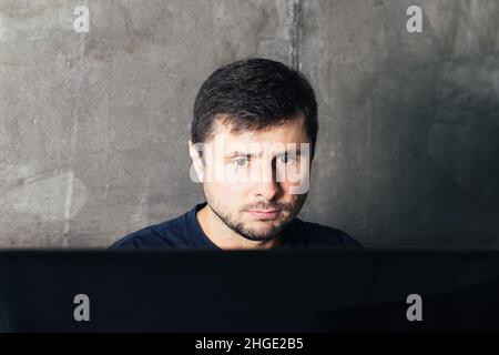 Handsome serious bearded man sitting at the office desk, working and looking at big computer screen against concrete wall in modern office Stock Photo