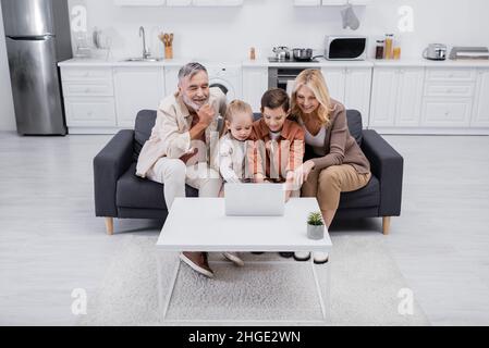 kids using laptop near happy grandparents on couch in kitchen Stock Photo