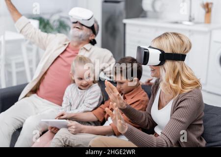 excited couple gaming in vr headsets near grandchildren using digital tablet on couch Stock Photo
