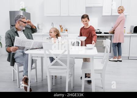 smiling man with newspaper looking at grandchildren setting table for breakfast Stock Photo