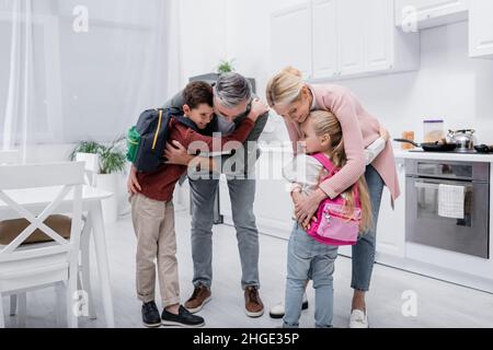 happy grandparents embracing children with backpacks in kitchen Stock Photo