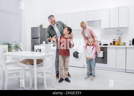 smiling grandparents putting backpacks on kids in kitchen Stock Photo