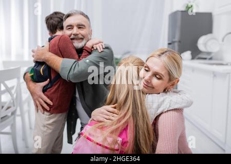 happy couple embracing grandchildren with backpacks in kitchen Stock Photo