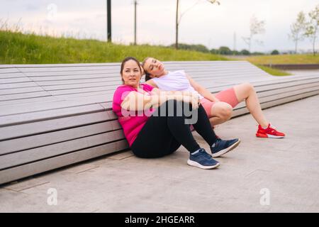 Wide shot of tired obese young woman resting by bench after intense exercise with personal trainer outdoor in city park. Stock Photo