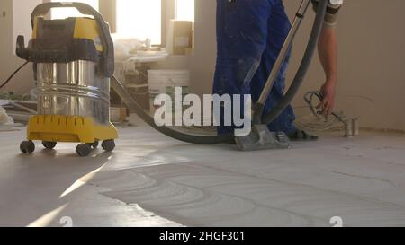 Workers in uniform cleaning room with a vacuum cleaner after installing wood parquet board during flooring work. Man vacuuming floor during repairs. Stock Photo