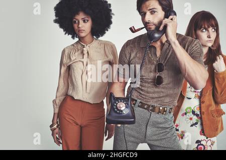Too busy for chicks. Two attractive young hippies standing behind a handsome man using a retro telephone. Stock Photo