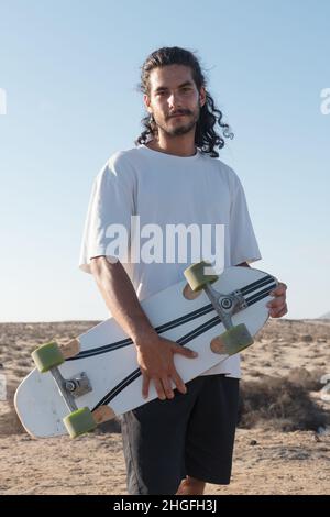 Young adult skater portrait standing outdoors holding his skateboard. He has long black hair and a beard. Stock Photo