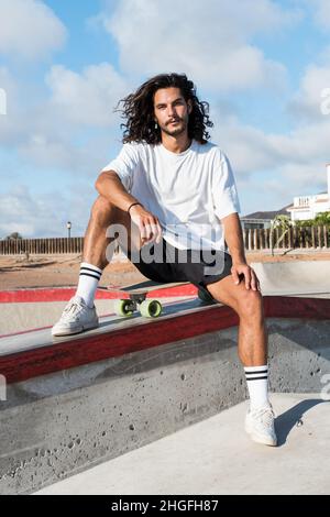 Young handsome skater sitting on his skateboard at the skate park. He is wearing summer clothes and he has long black hair and a beard. Stock Photo