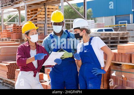Managers and workers in protective mask are discussing work plan on site of an open-air construction store Stock Photo