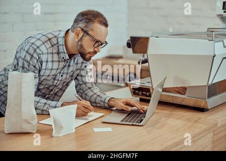 Male cafe owner using laptop and writing in notebook Stock Photo