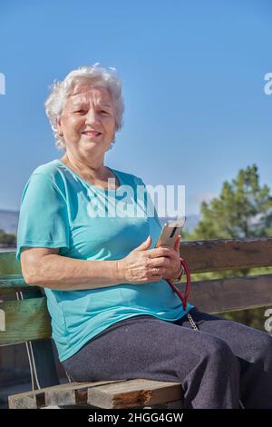 An optimistic old lady sitting on a bench using a cell phone, under a cloudless blue sky on a sunny day and looking at the camera Stock Photo