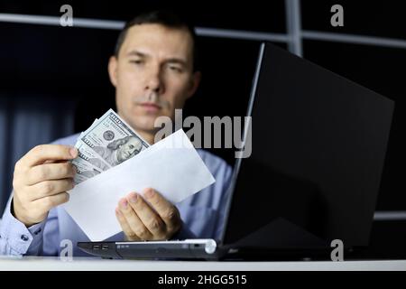 US dollars in male hands. Man in office clothes pulls money out of an envelope sitting at laptop, wages, bonus or bribe concept Stock Photo