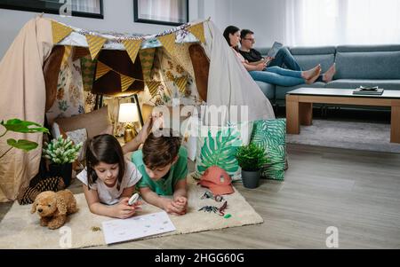 Children playing camping at home while their parents read and look at the mobile sitting on the sofa Stock Photo