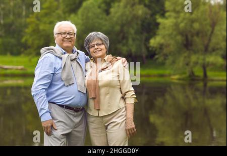 Portrait of smiling elderly pensioners standing in the park by the lake in summer. Stock Photo