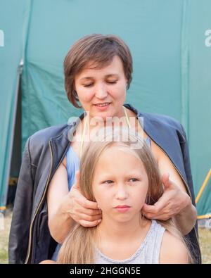 Mother styling long blond hair to her daughter in camping over of tent background. Stock Photo