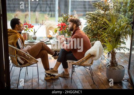 Two multiracial male friends having close conversation, sitting by a dinner table at backyard Stock Photo