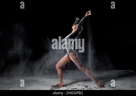 gorgeous slender ballet dancer woman wearing bodysuit, posing dancing among the cloud of flying flour on black studio background, make performance. Ar Stock Photo