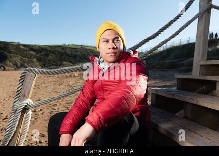 Portrait of young latin man wearing winter clothes and sitting on stairs Stock Photo