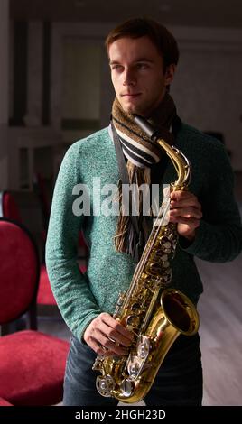 Young guy music artist jazzman holding a saxophone in his hands. The dark room of the concert hall, illuminated by the light from the window Stock Photo