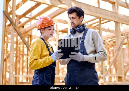 Two man and woman in uniform and helmet explore construction documentation on the building site near the wooden building constructions, have conversat Stock Photo