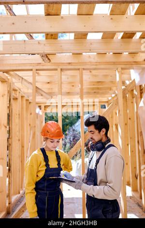 Two caucasian civil engineers dressed in uniform explore construction documentation on the building site near the wooden building constructions, weari Stock Photo