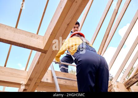 two contractors in working clothes uniform climb to the roof of wooden building house on stepladder, rear view on female climbing to height, outdoors. Stock Photo