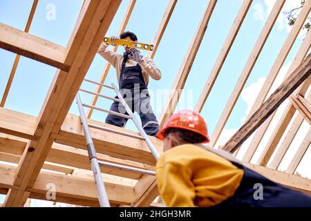 two contractors in working clothes uniform climb to the roof of wooden building house on stepladder, rear view on female climbing to height, outdoors. Stock Photo